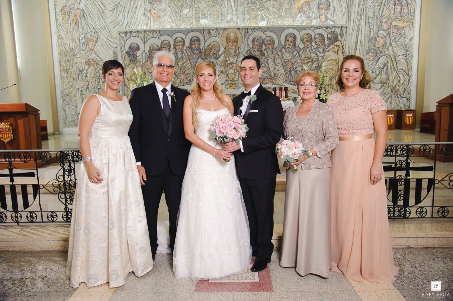 Wedding party posing in church, bride in white gown holding pink bouquet, flanked by family in formal attire.