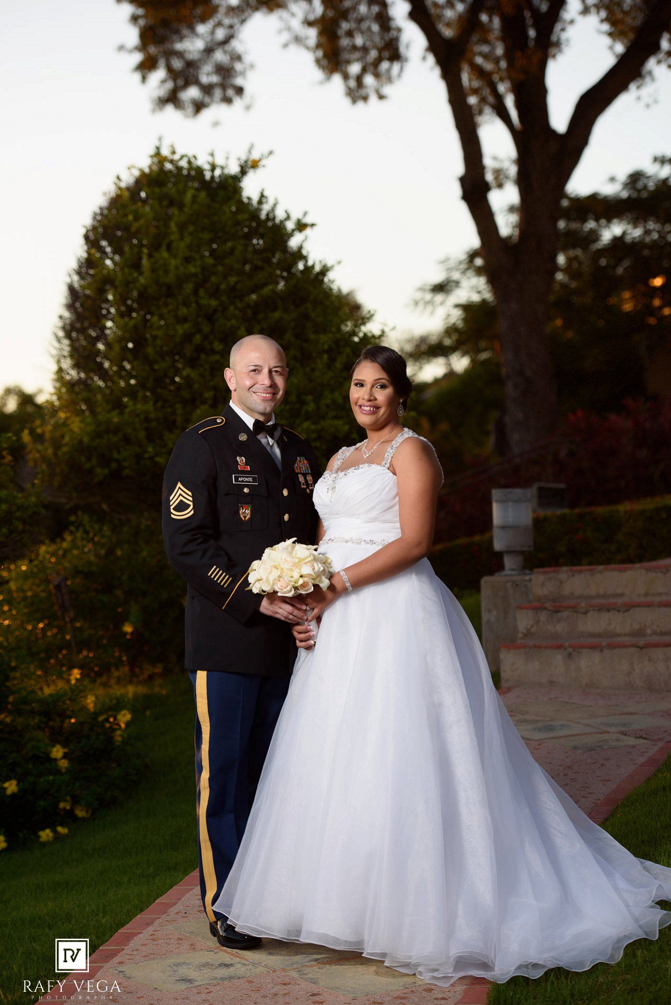 Military groom in Army dress uniform stands with bride holding white bouquet in ball gown at outdoor wedding.