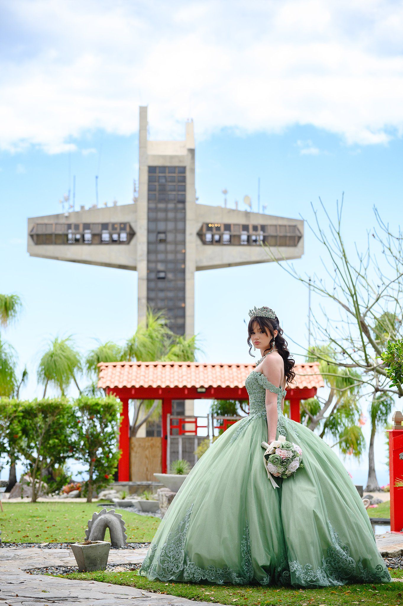 Elegant quinceañera in sage green ball gown with tiara holds bouquet before large cross monument outdoors.