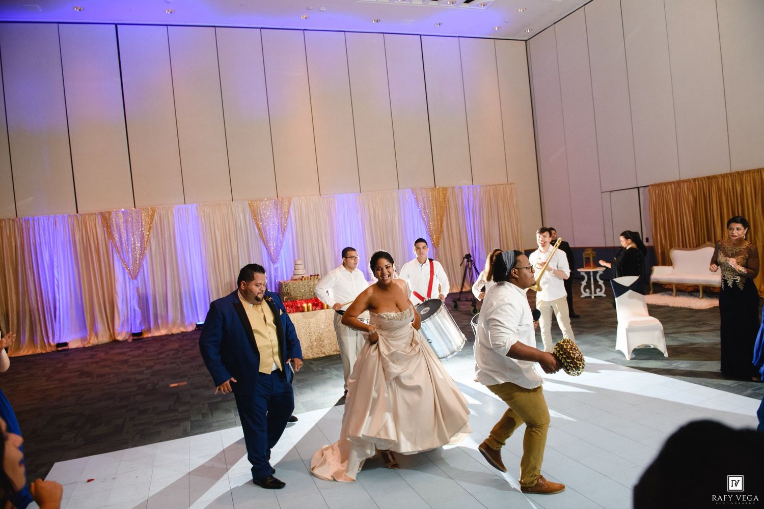 Bride in white gown dancing with guests at elegant wedding reception venue with draped backdrop lighting.