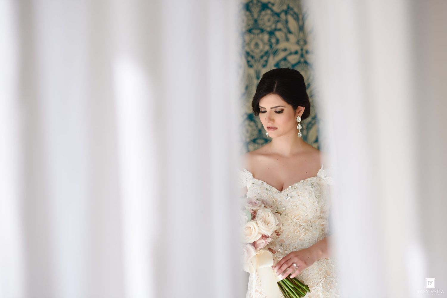 Elegant bride in lace off-shoulder gown holding pink bouquet, framed by white curtains in soft natural light.