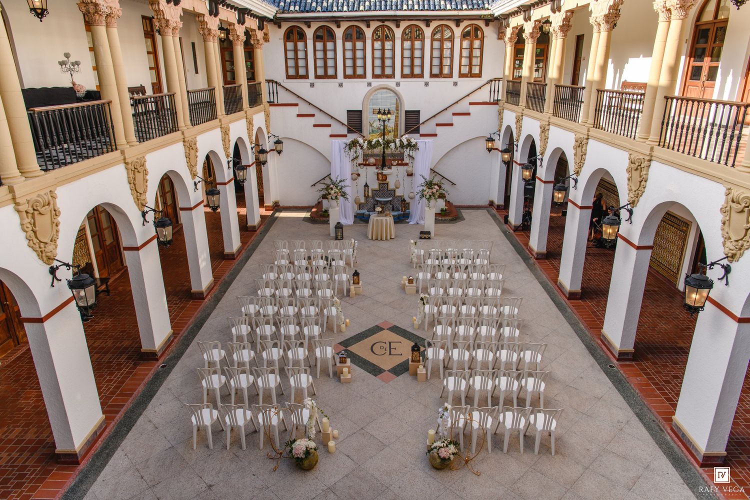Elegant outdoor courtyard wedding setup with white chairs arranged in rows under a grand colonial-style building with arched balconies.