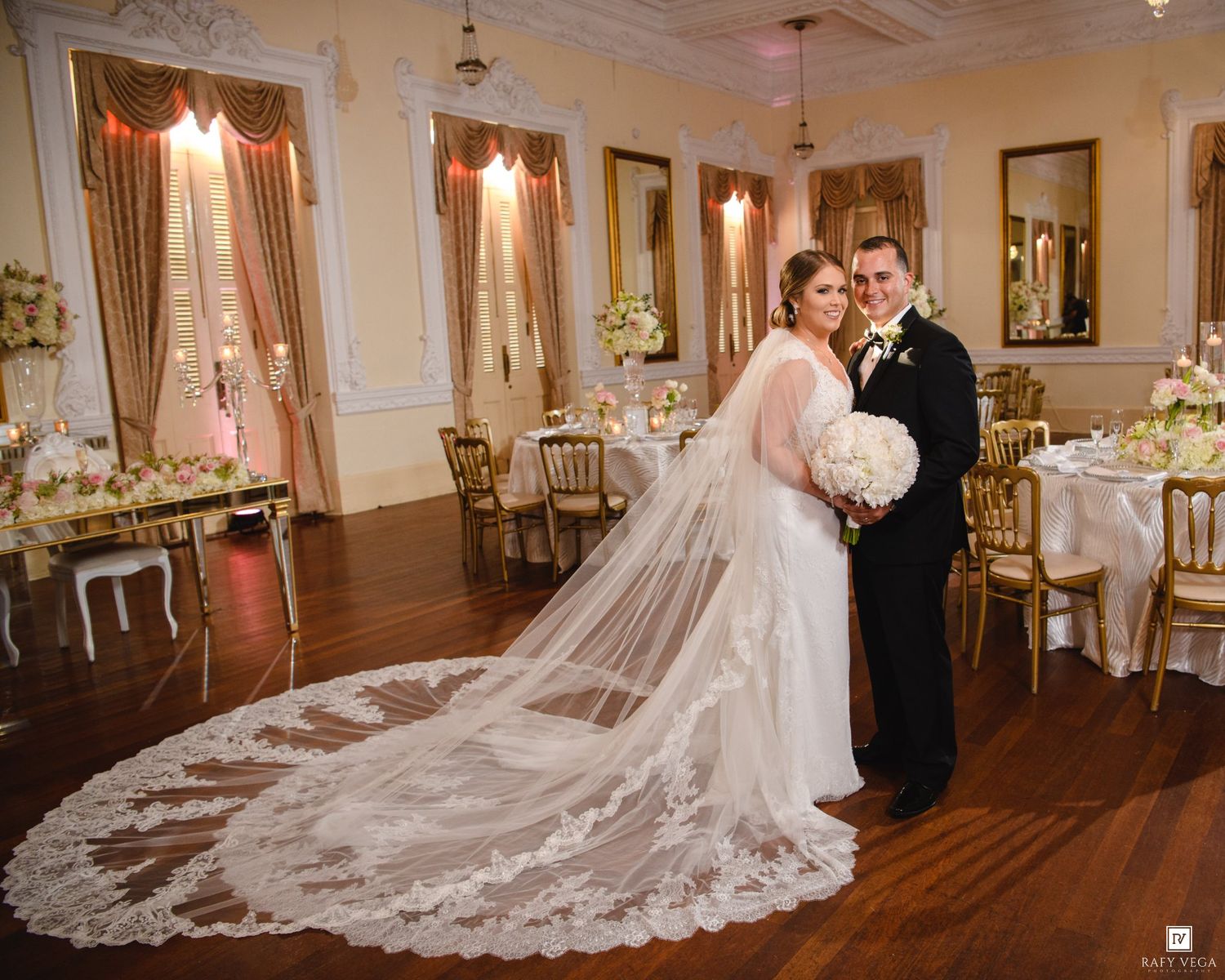 Bride and groom pose in elegant ballroom with gold chairs, floral centerpieces, and bride wearing long lace cathedral veil.