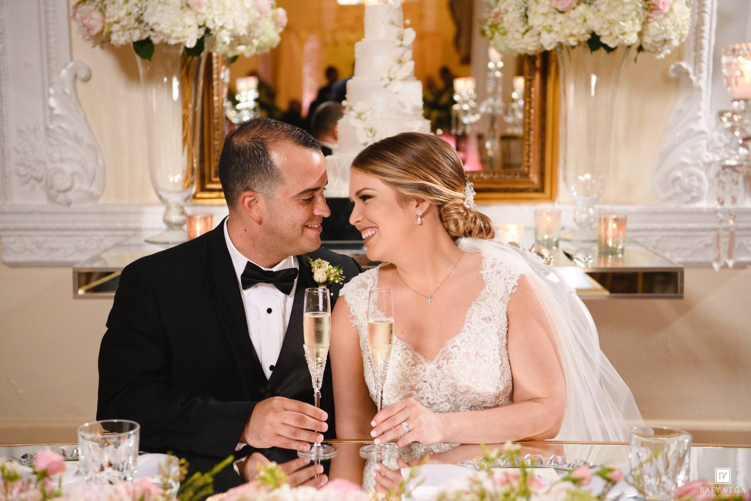 Bride and groom share a romantic toast with champagne flutes at their wedding reception sweetheart table with floral decor.