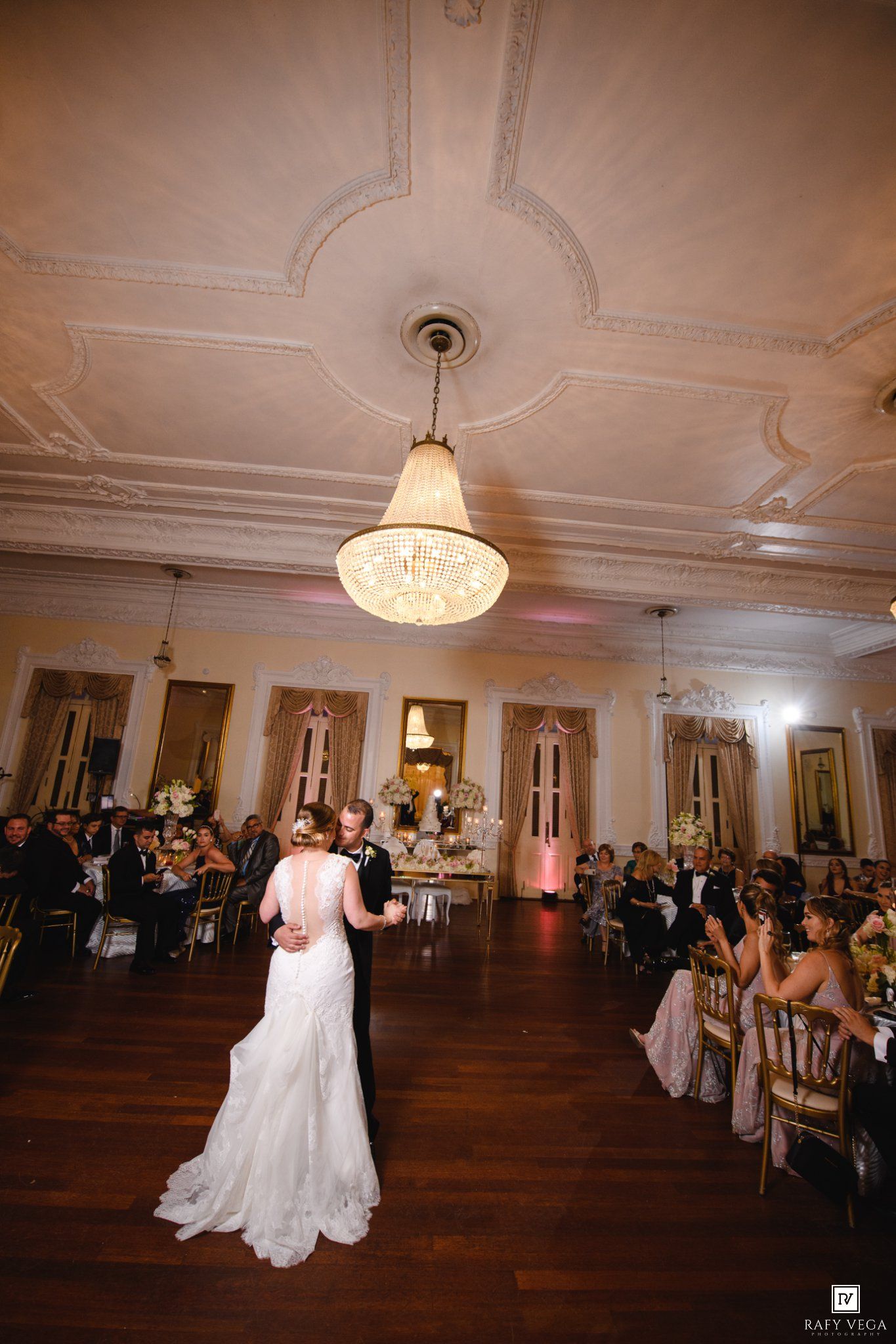 Bride and groom share their first dance under an elegant chandelier in a grand ballroom filled with wedding guests.