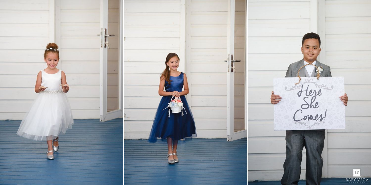 Three children in wedding attire, two flower girls in white and navy dresses and a ring bearer holding a Here She Comes sign.