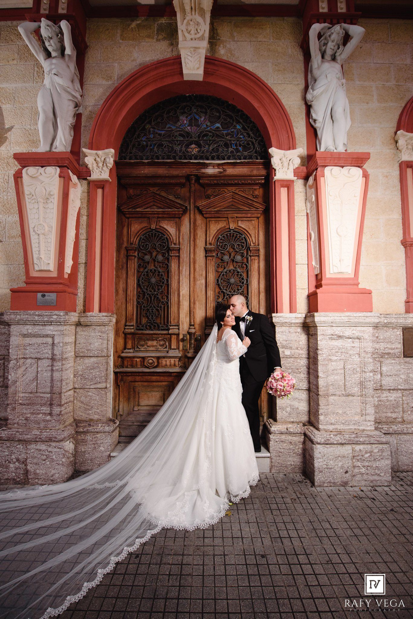 Groom kissing bride beside grand arched doorway with sculpted figures at wedding venue.