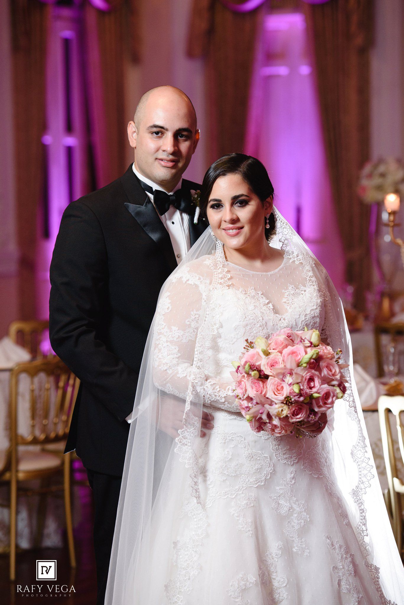 Bride in lace gown holding pink bouquet poses with tuxedo-clad groom in elegant pink-lit wedding reception venue.