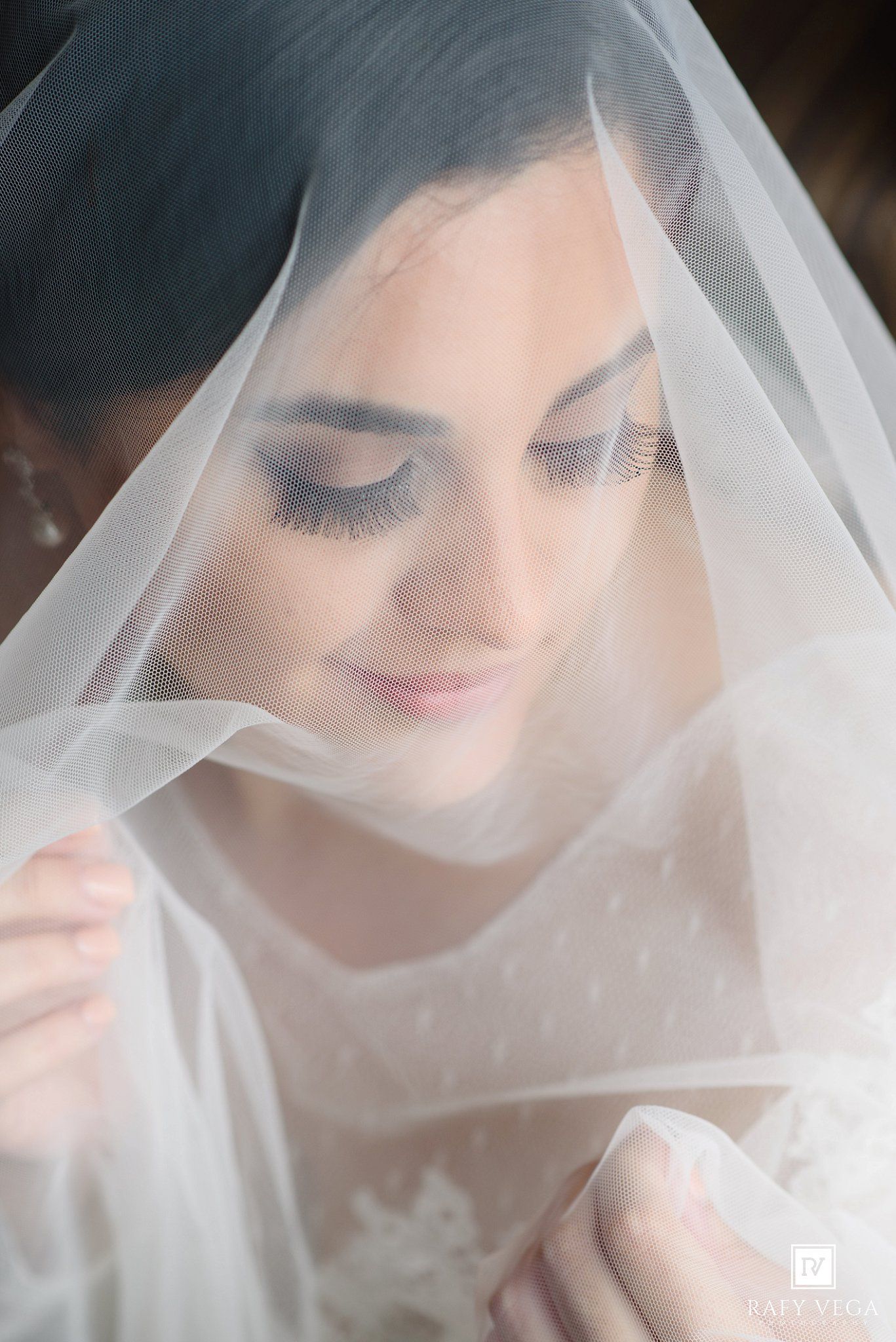 Bride draped in a sheer white veil, showcasing elegant makeup with long lashes and a serene expression.