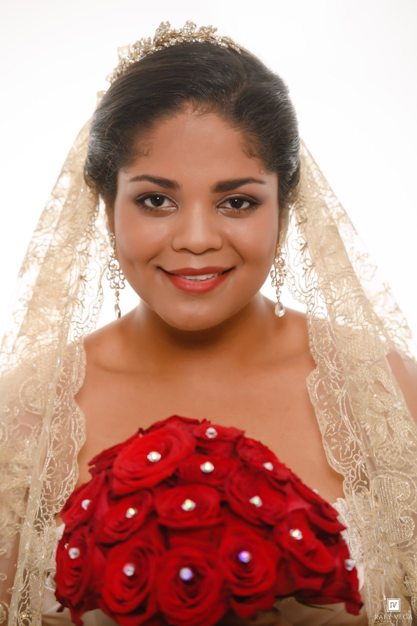 Bride wearing a gold lace veil and tiara holds a red rose bouquet with pearl accents, smiling elegantly.