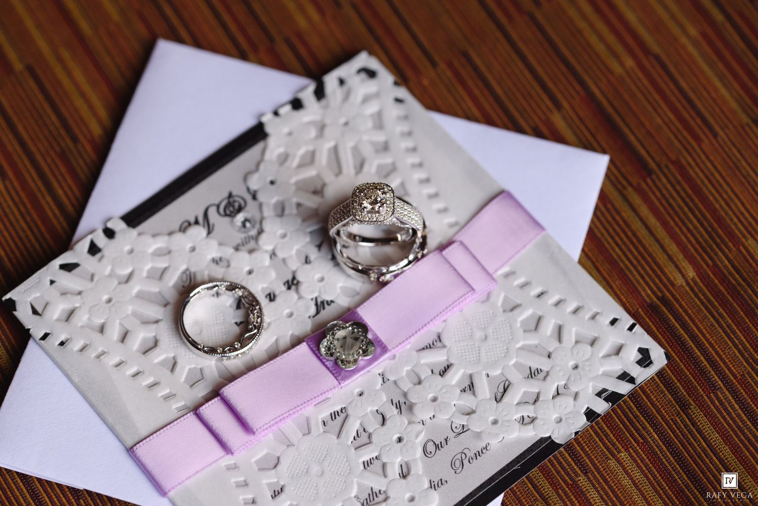 Elegant wedding invitation with lace design, pink ribbon bow, and two silver rings displayed on a wooden surface.