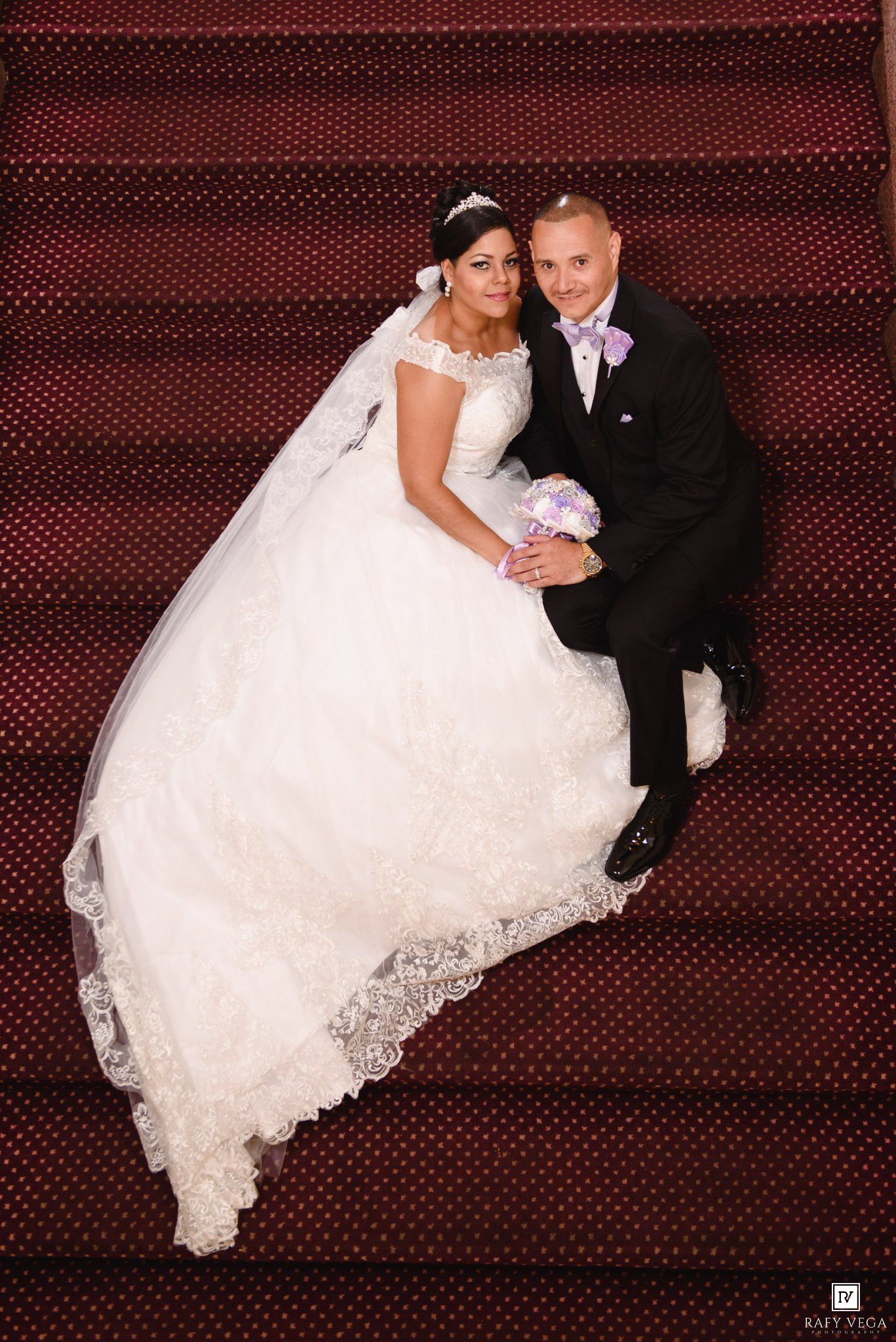 Bride in white ball gown and groom in black suit sit on red carpeted stairs, smiling, holding purple bouquet.