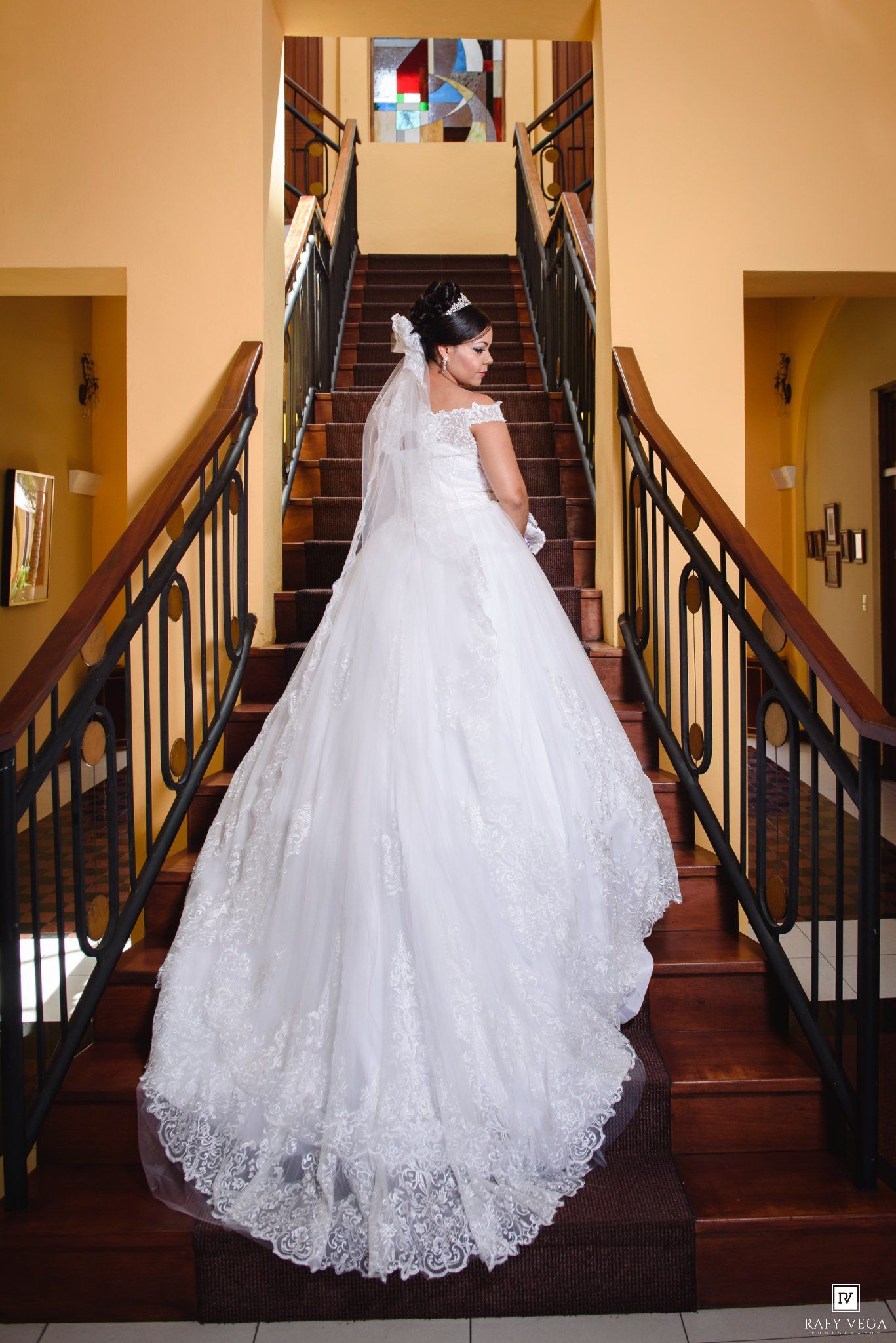 Bride in elegant white ball gown with lace train stands on grand staircase with stained glass window backdrop.