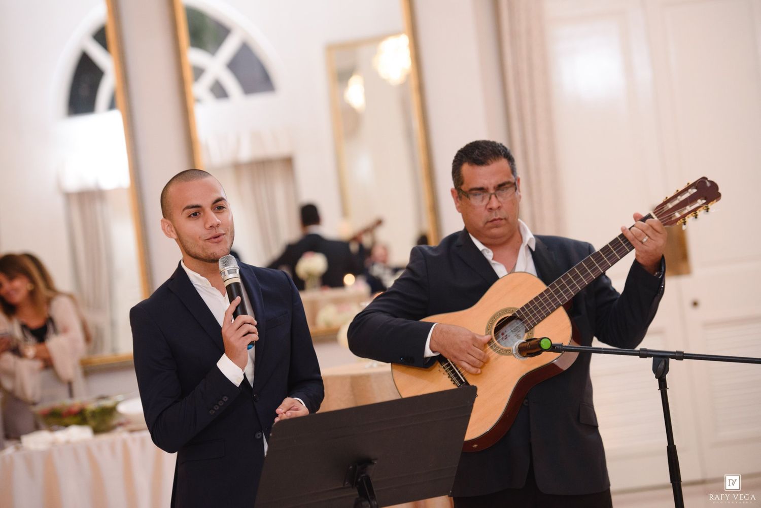 Two men in suits perform at an elegant event, one singing into a microphone and one playing acoustic guitar.