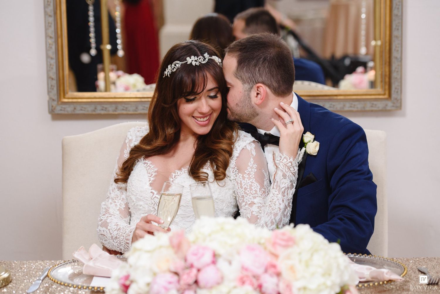 Bride and groom share a romantic kiss at wedding reception sweetheart table with champagne and floral centerpiece.
