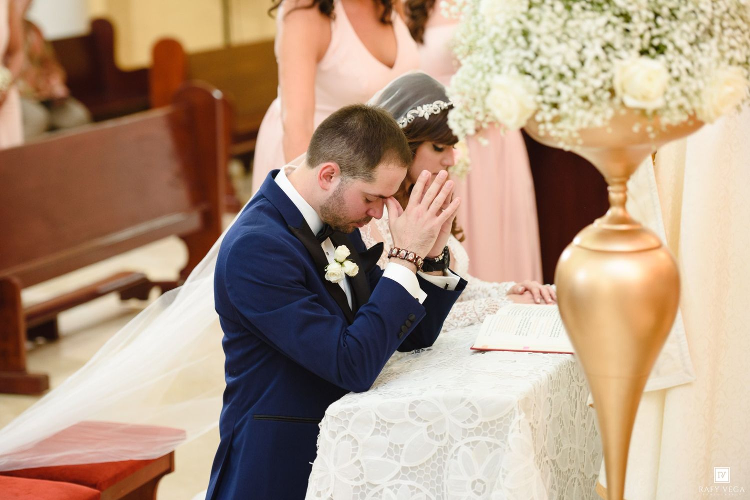 Bride and groom kneeling in prayer at church altar during wedding ceremony, with bridesmaid in pink dress behind them.