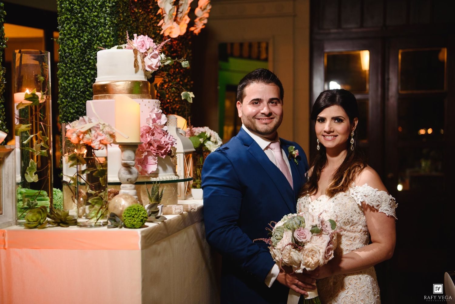 Bride and groom smiling beside elegant multi-tier wedding cake adorned with pink flowers at reception.