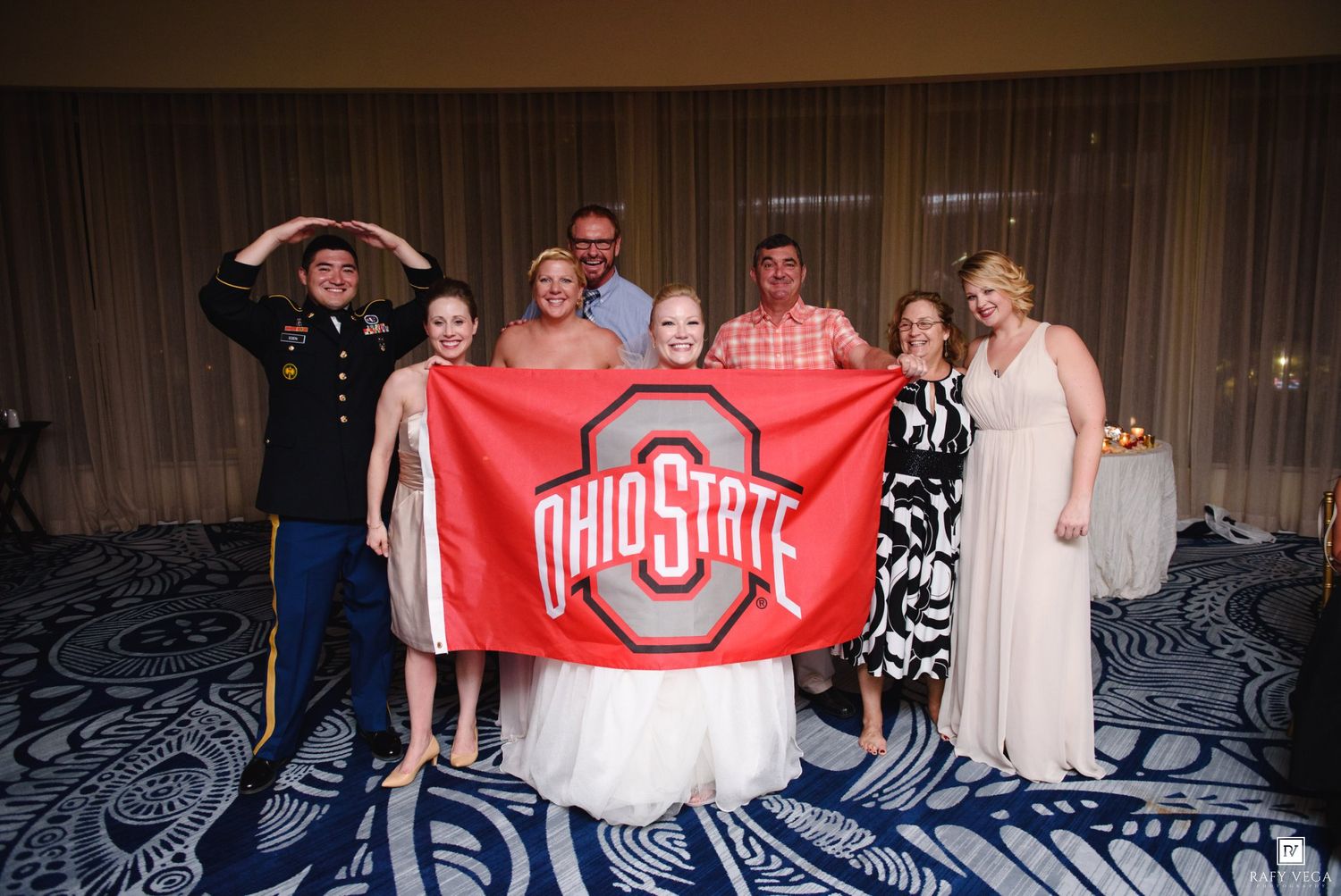 Wedding group photo with Ohio State flag, featuring bride, groom, and guests celebrating at indoor reception.