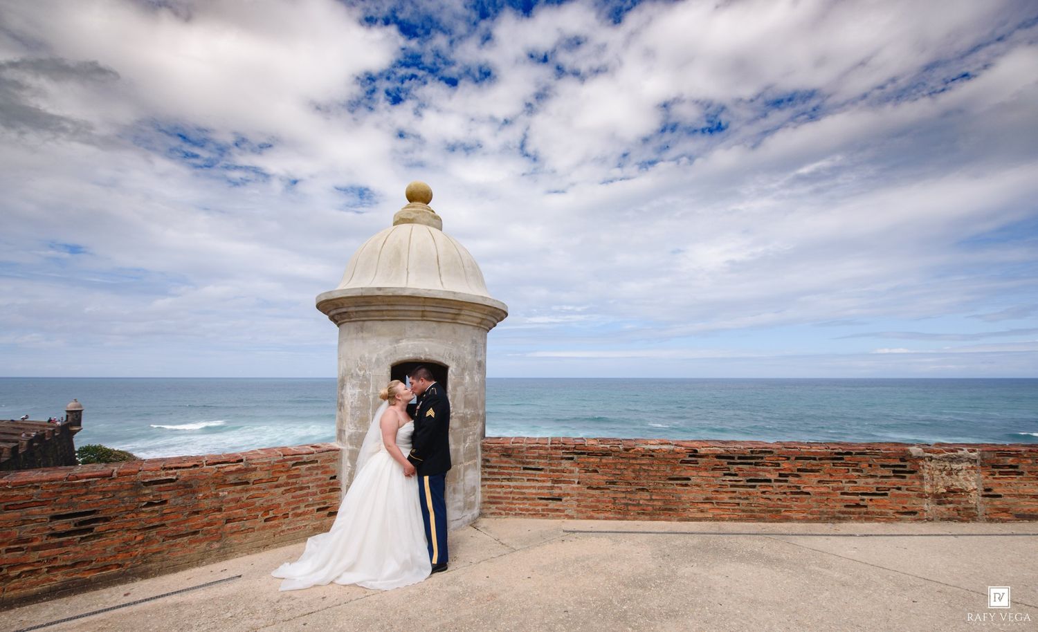 Bride and groom embrace by a historic stone turret at El Morro fort with ocean views in Puerto Rico.