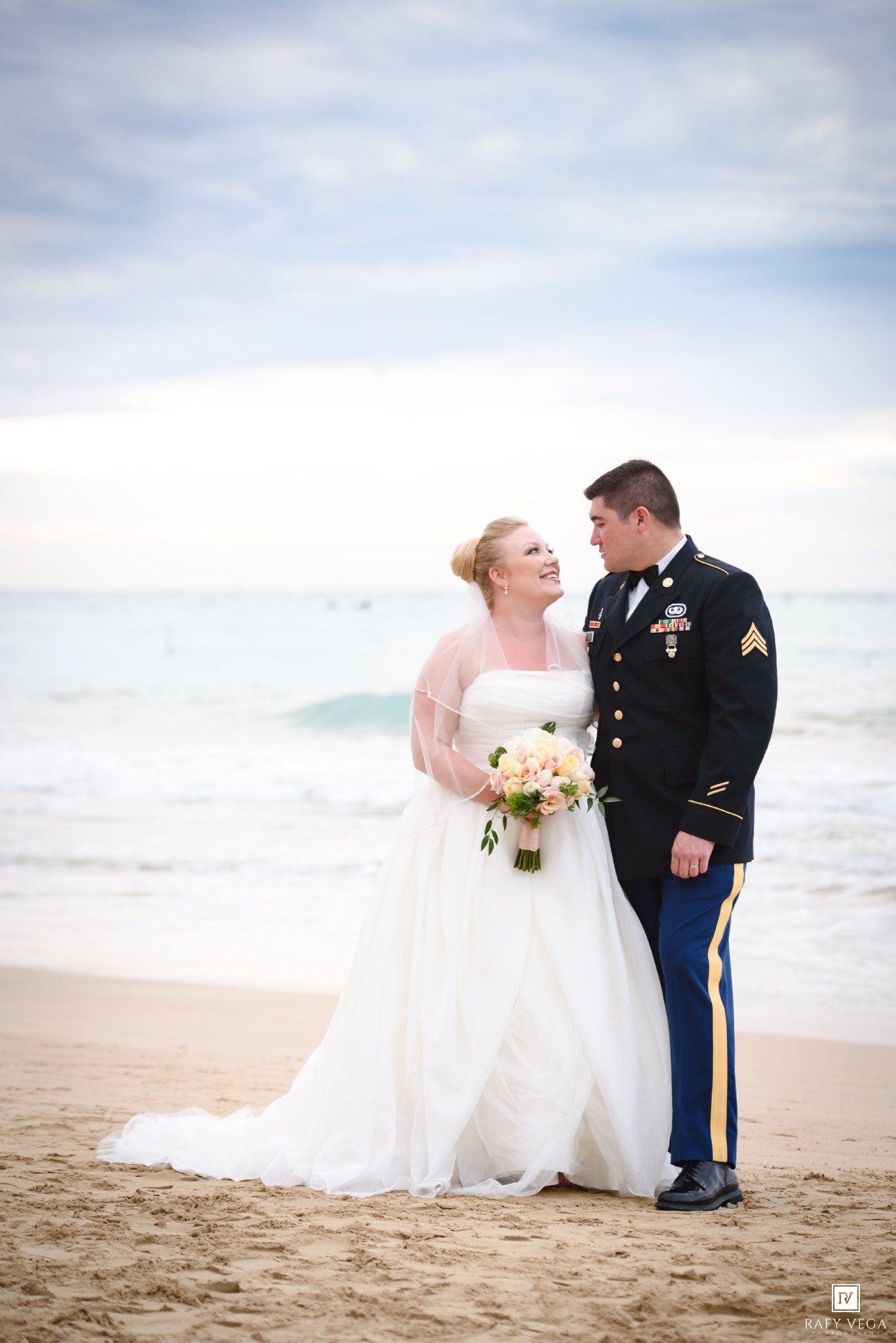 Bride in white gown and groom in military dress uniform share a romantic moment on a sandy beach.
