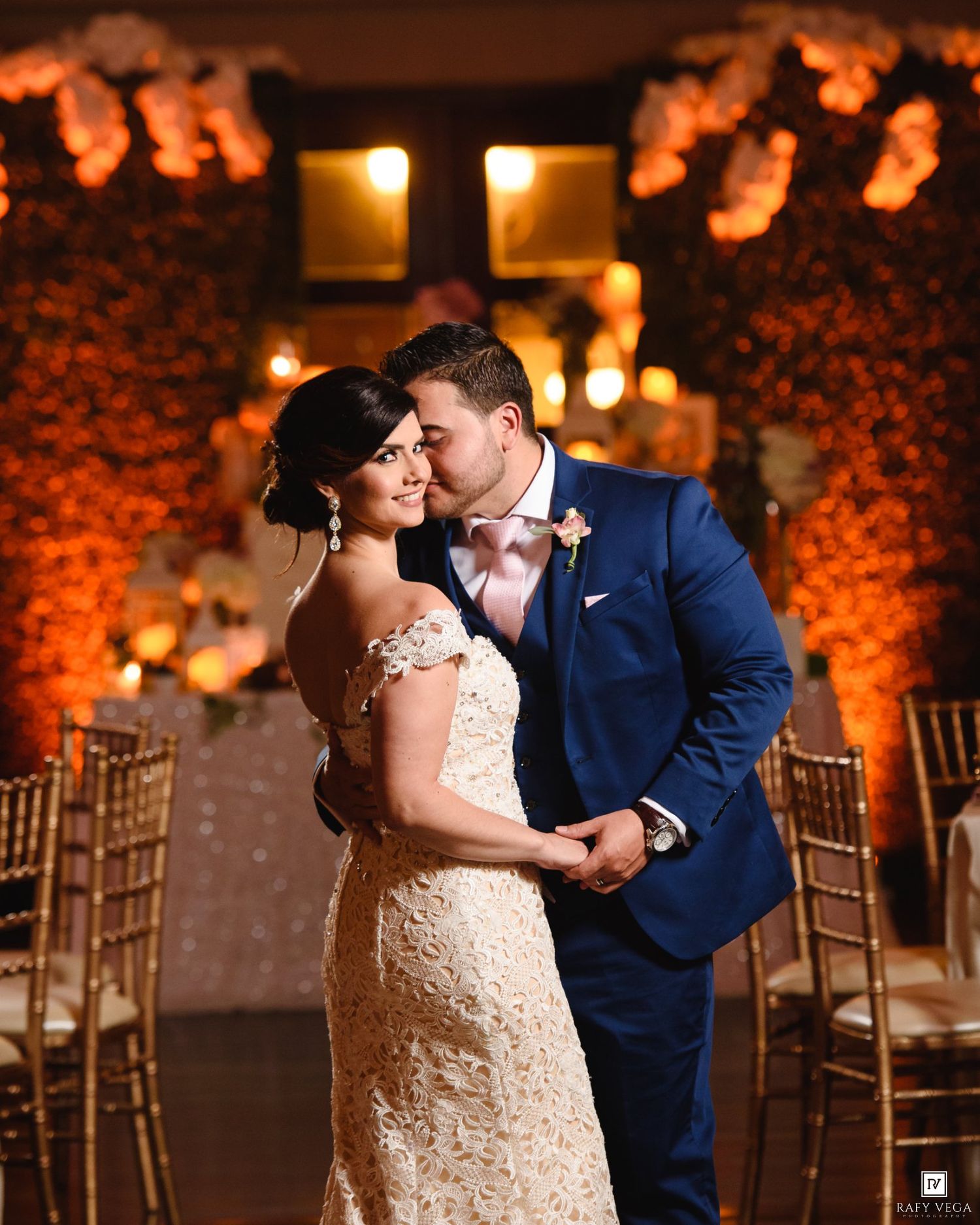Bride and groom share a romantic embrace at elegant wedding reception with warm golden lighting and floral decor.