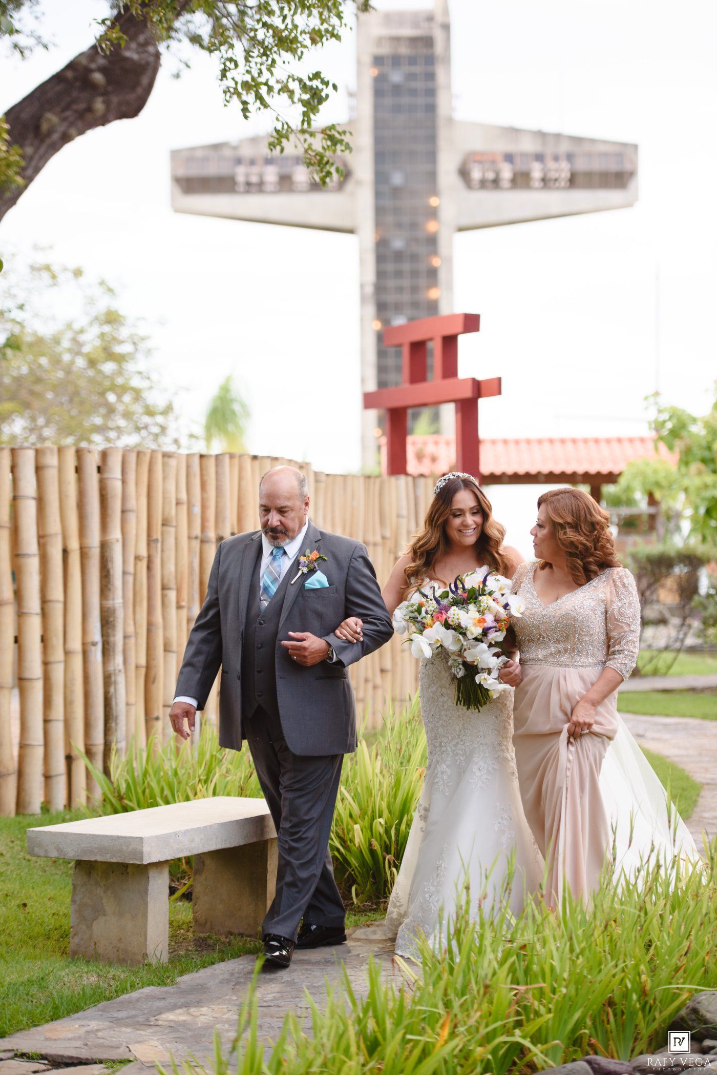 Bride walks with parents toward church entrance, holding colorful bouquet, with bamboo fence and cross in background.