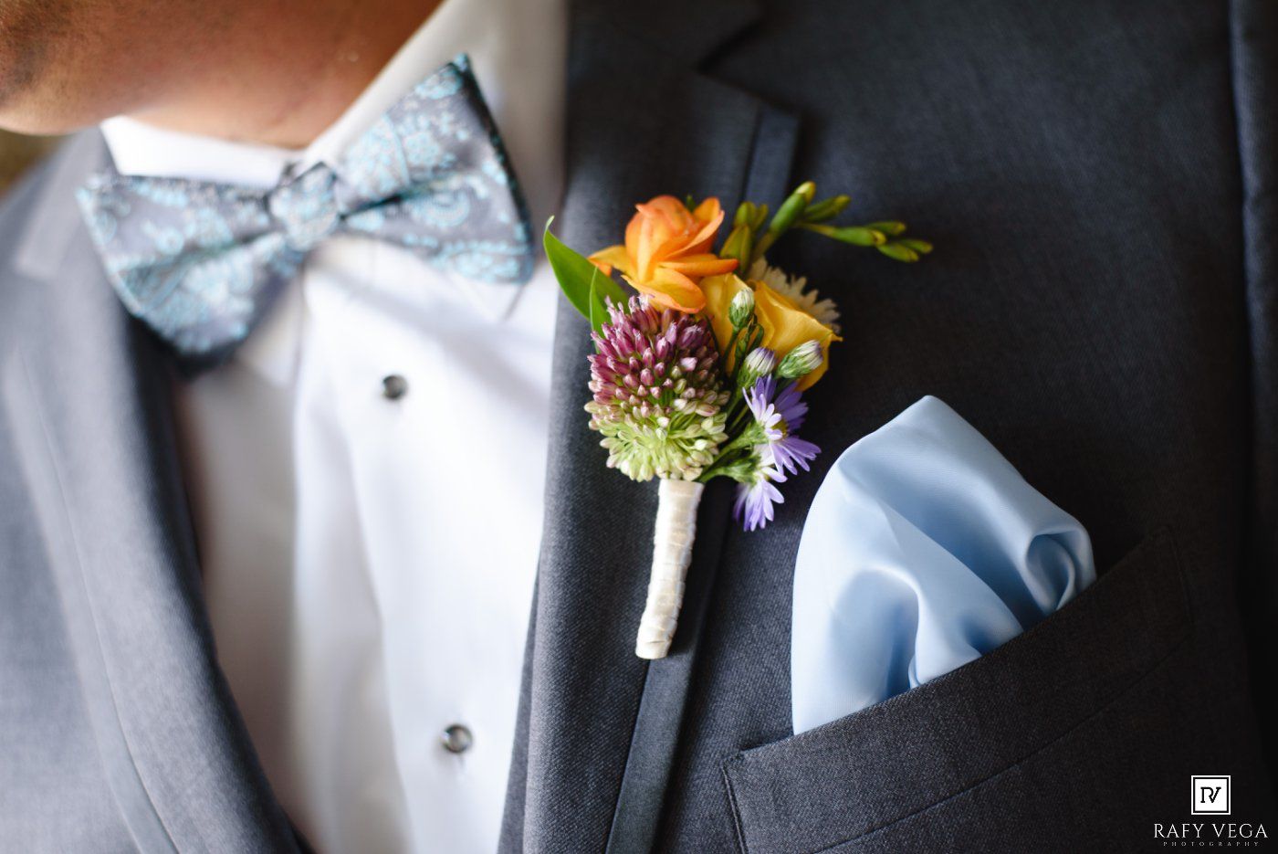 Groom wearing gray suit with colorful boutonniere, light blue bow tie, and matching pocket square at wedding.