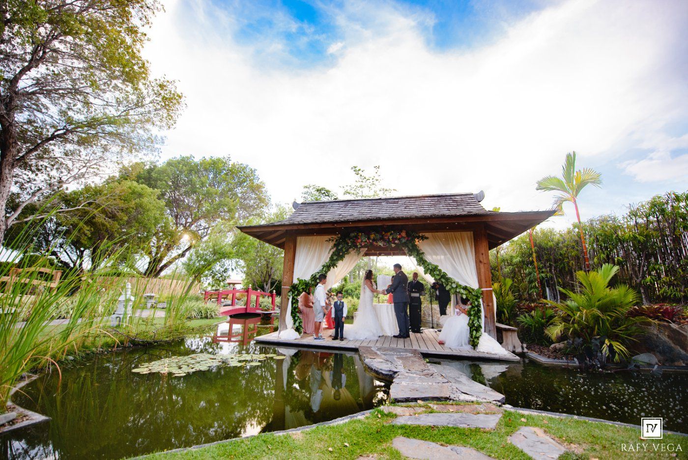 Elegant outdoor wedding ceremony at a gazebo beside a tranquil pond with lush tropical gardens.