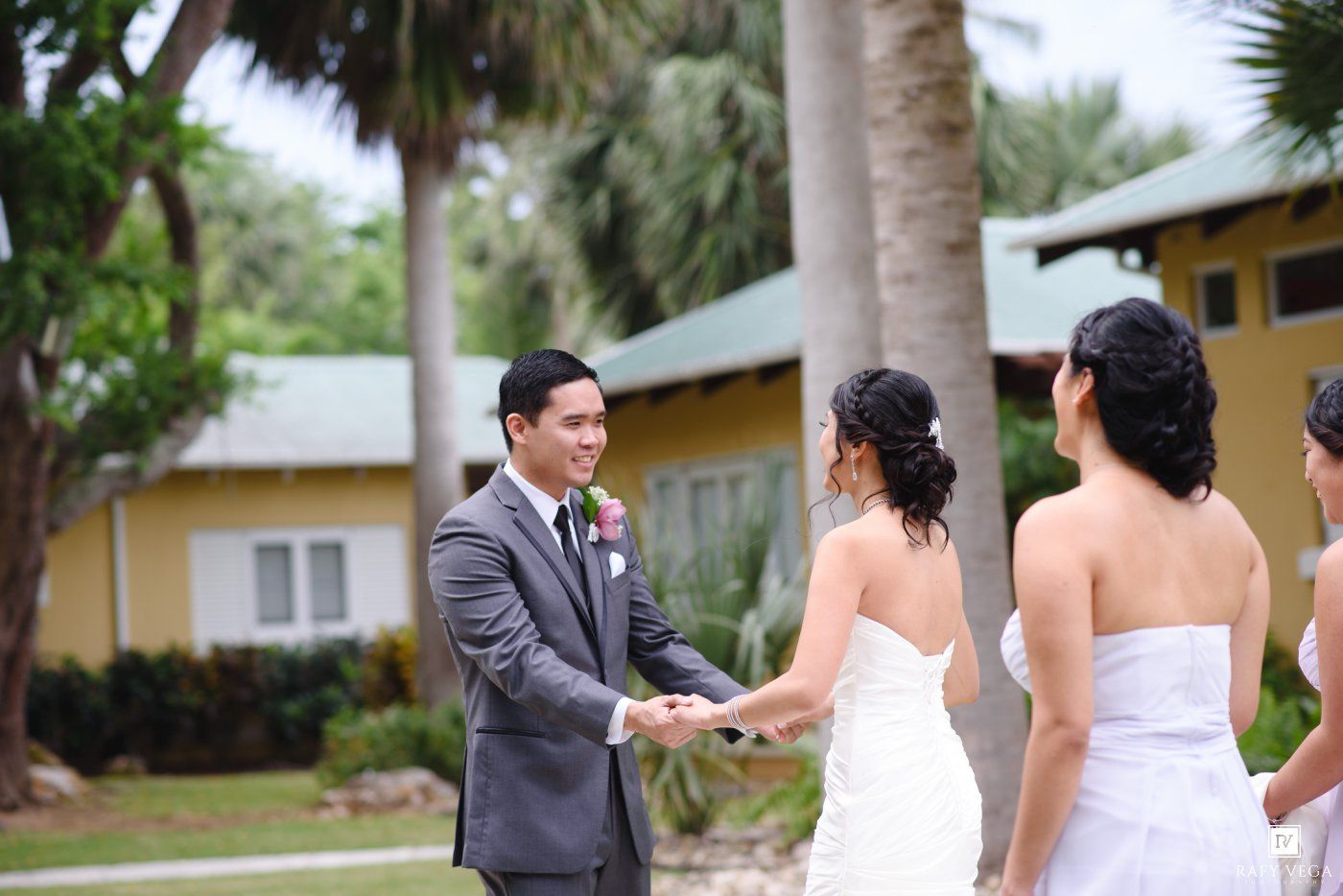 Groom in gray suit holds hands with bride in white dress during outdoor wedding ceremony near palm trees.