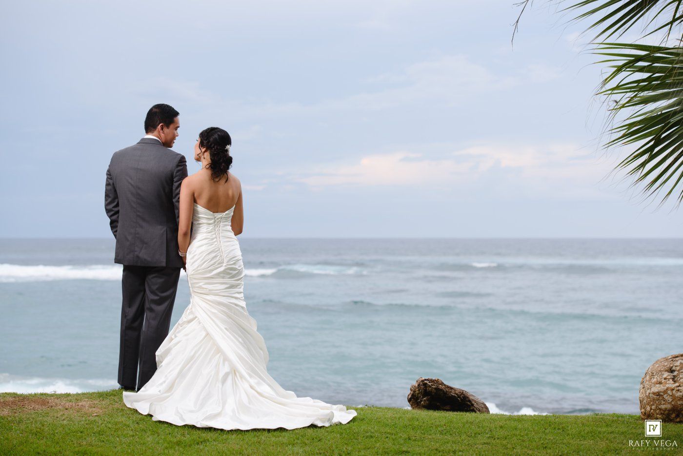 Bride and groom stand together on a lush green lawn overlooking a serene ocean view on their wedding day.