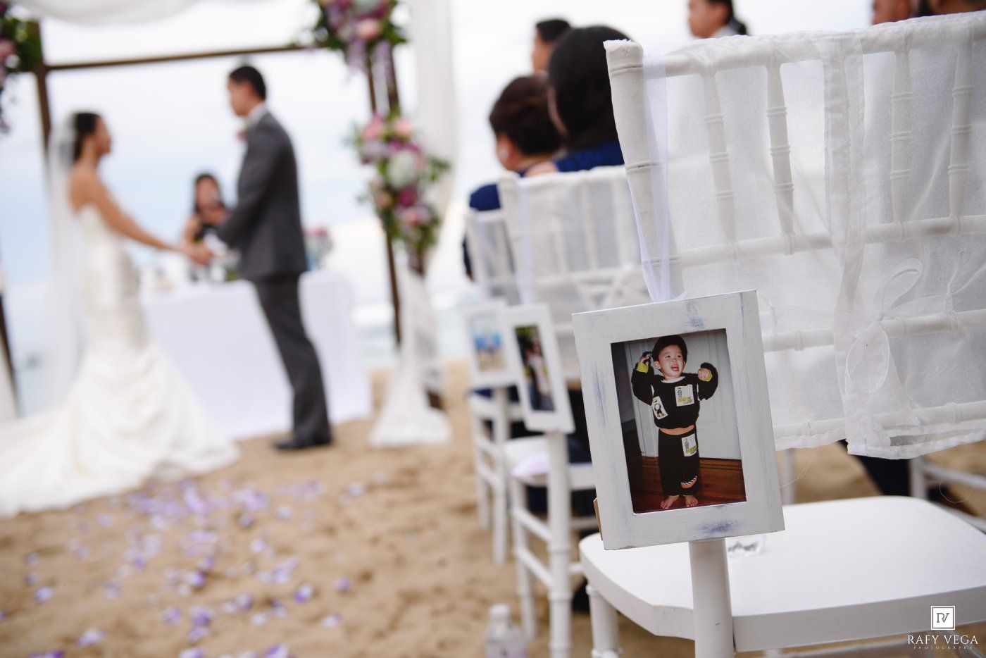 Beach wedding ceremony with bride and groom at altar, white chairs lining the aisle, and a framed photo displayed on a chair.