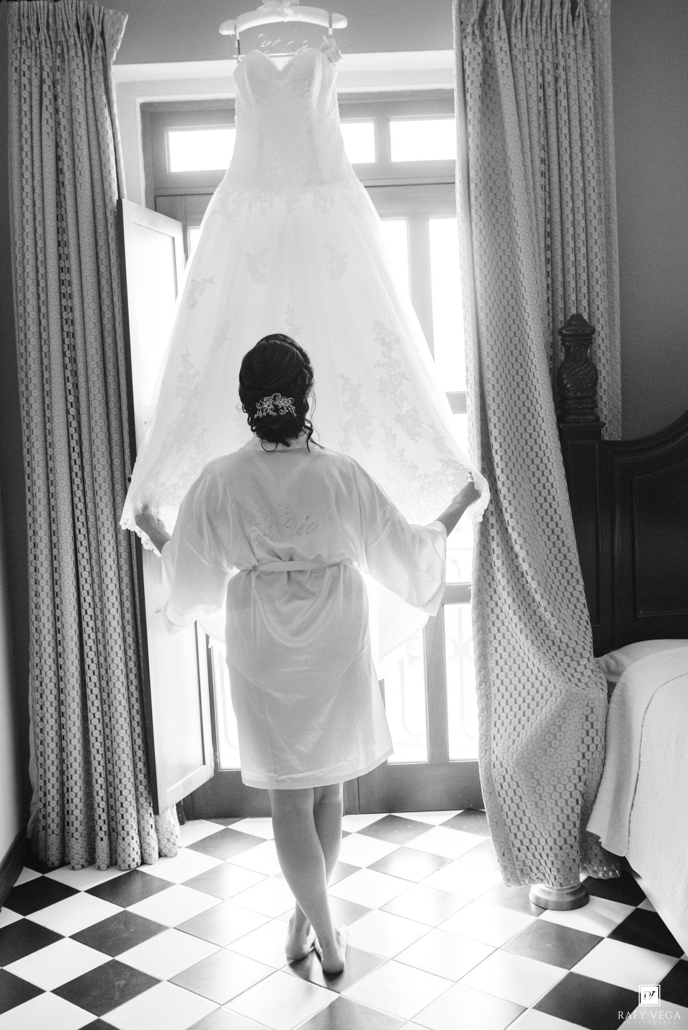 Bride in white robe admiring her wedding dress hanging by sunlit windows on checkered floor.