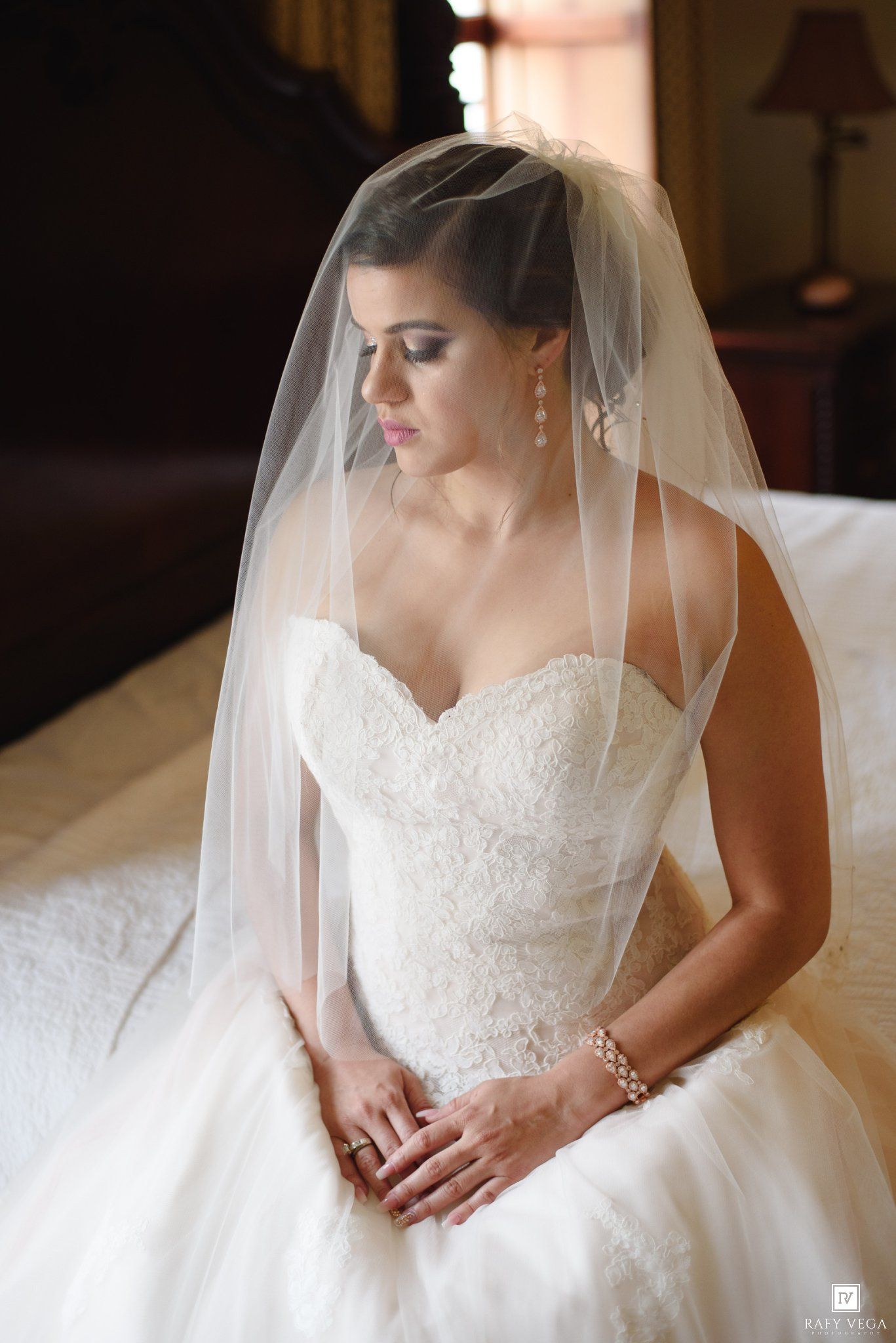 Bride in strapless lace wedding gown and veil sits gracefully, wearing pearl bracelet and earrings in soft natural light.