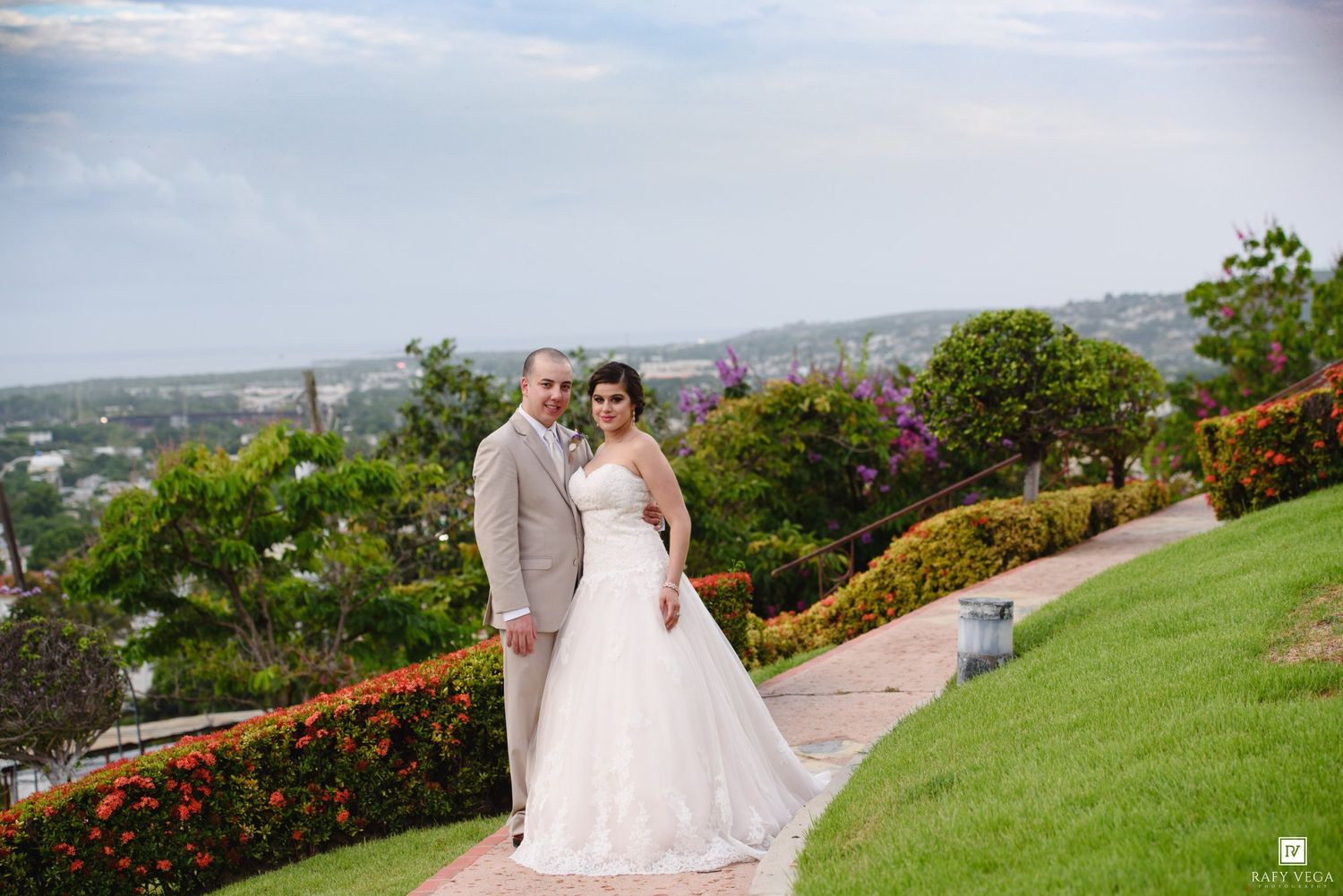 Wedding couple poses together on a lush green garden path with a panoramic coastal city view behind them.