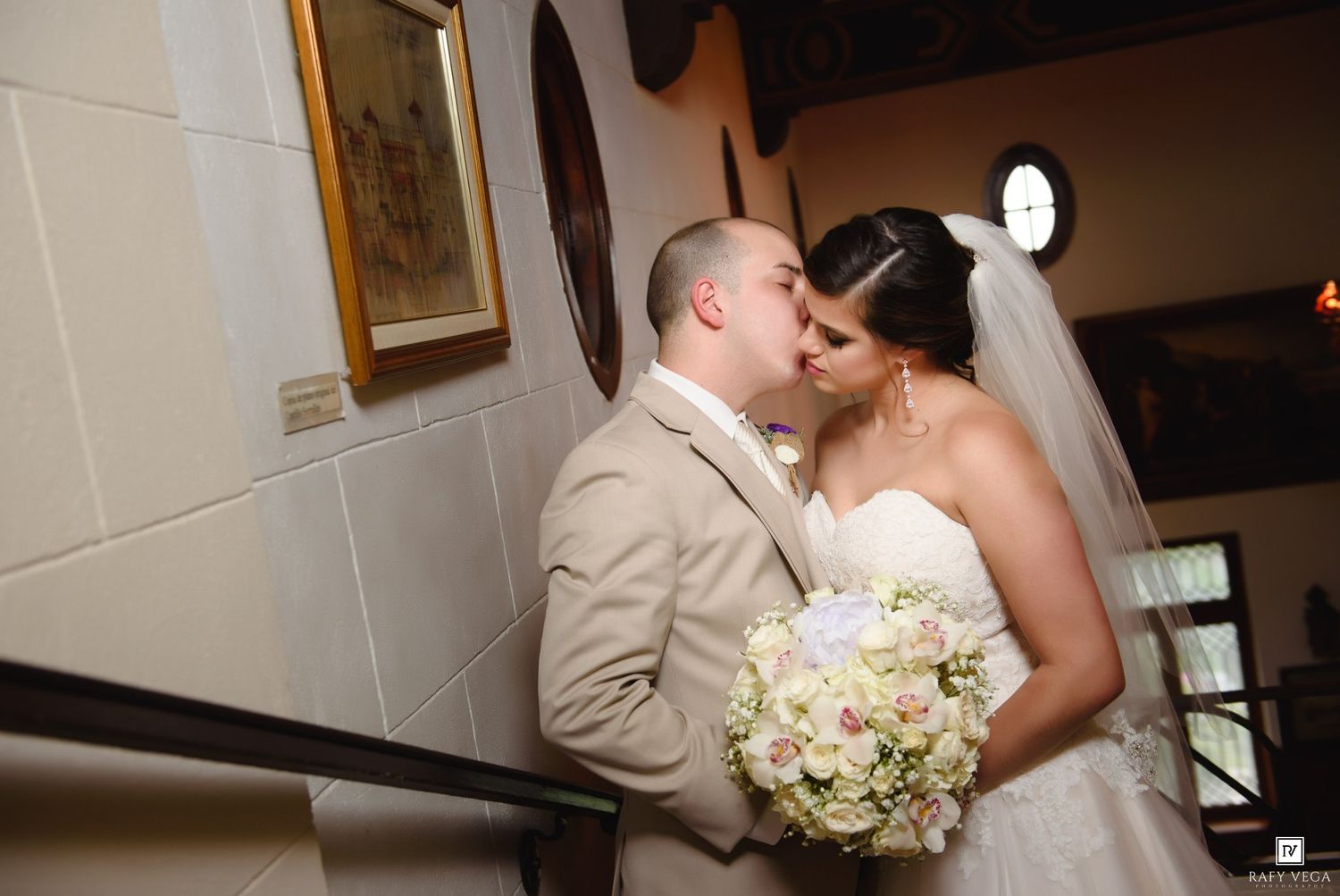 Bride and groom share a romantic kiss on a staircase, bride holding a white floral bouquet in an elegant wedding venue.