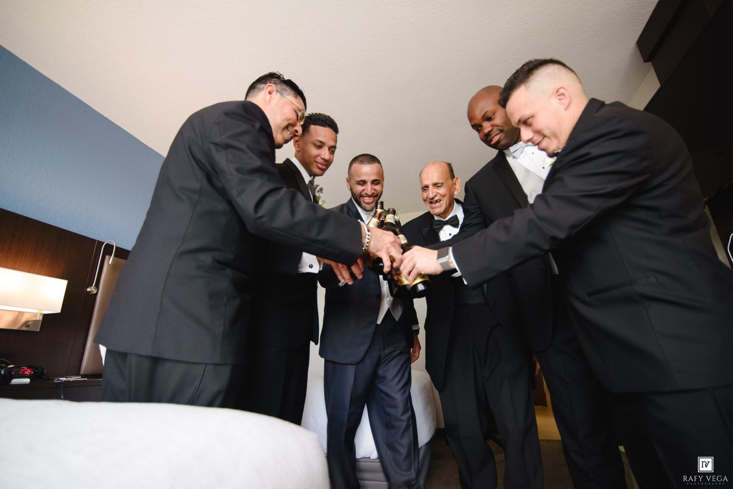 Six men in formal black suits gather in a circle toasting with champagne bottles at a wedding celebration.