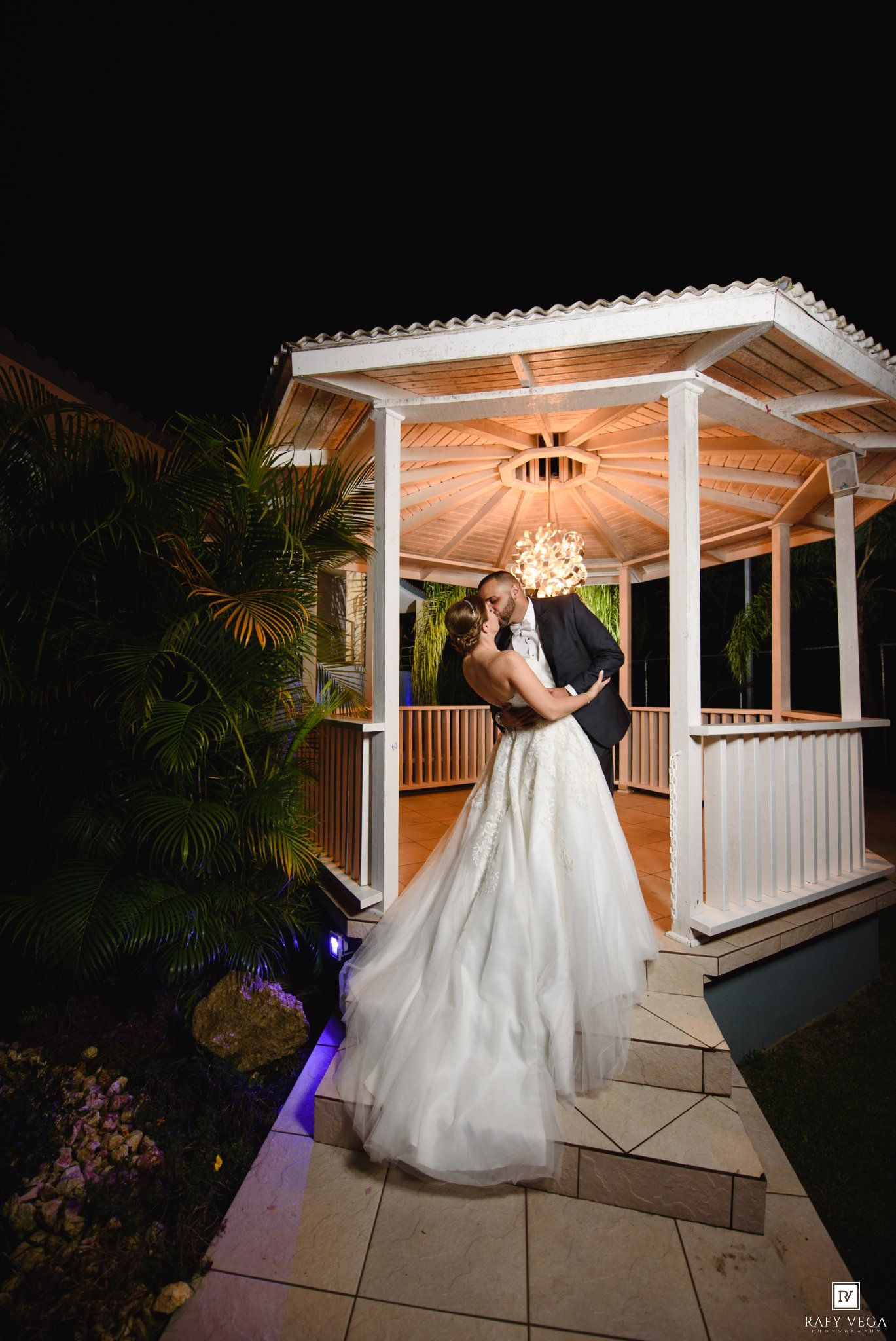 Bride and groom share a romantic kiss under a lit gazebo at night, surrounded by lush tropical greenery.