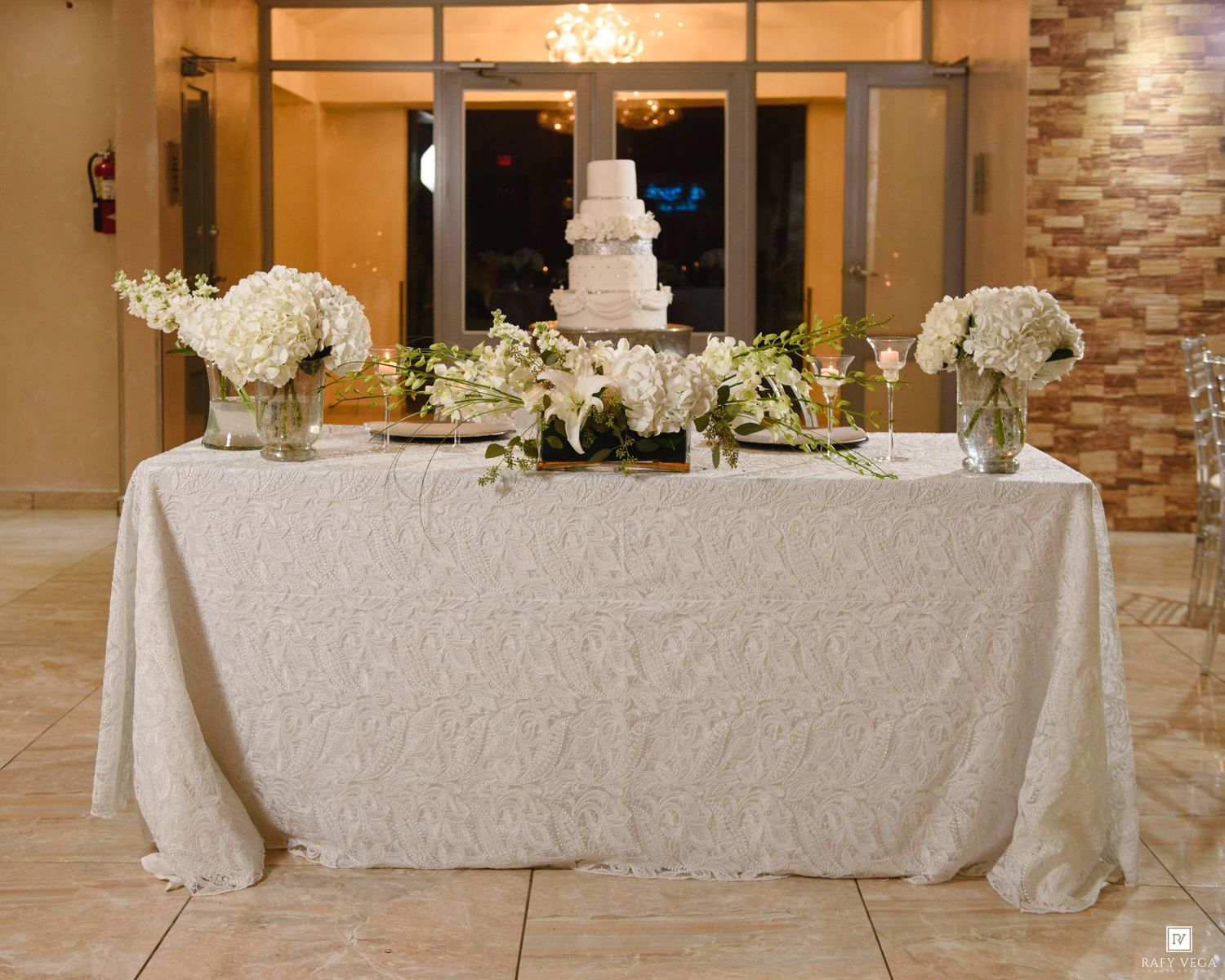 Elegant wedding cake table with white floral arrangements and lace tablecloth in a warmly lit reception venue.