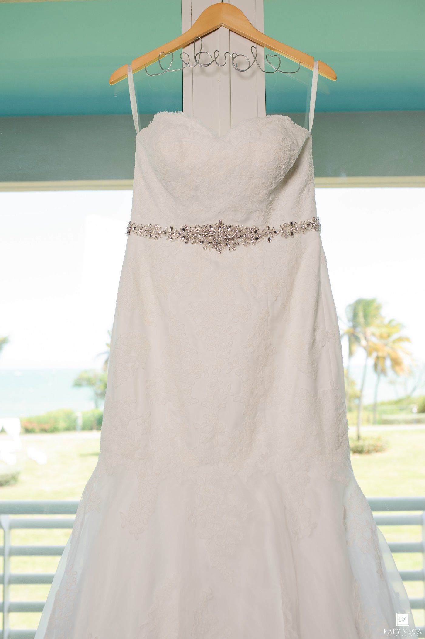 White strapless mermaid wedding gown with crystal belt hanging on decorative hanger near window with tropical ocean view.