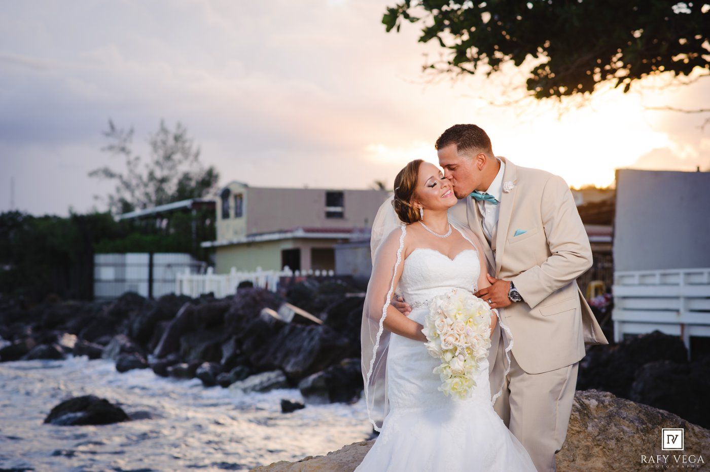 Bride and groom share a romantic kiss at sunset near rocky shoreline, bride in white gown with veil holding bouquet.