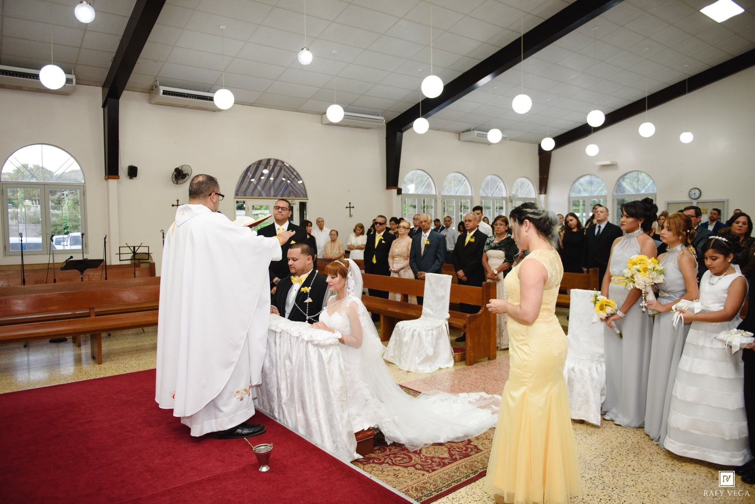 Priest officiating wedding ceremony with bride and groom kneeling at altar in decorated church with bridesmaids.