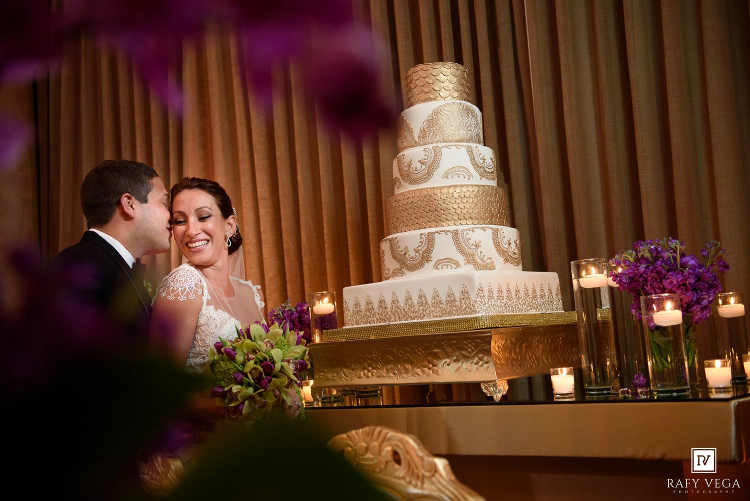 Bride and groom smiling beside a tall elegant white and gold wedding cake with candles and purple flowers.