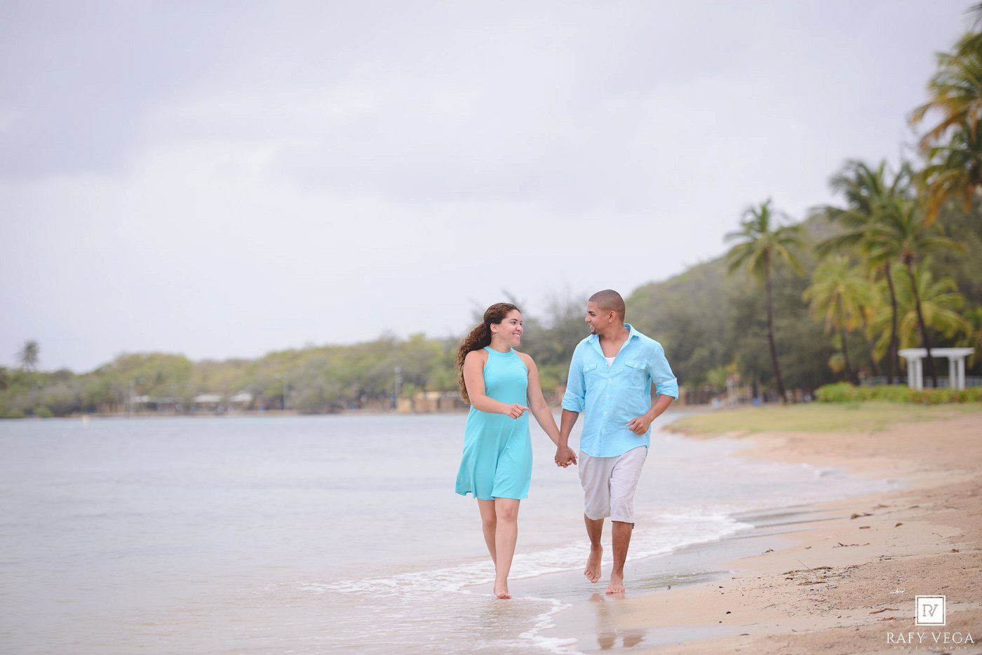 A romantic couple walks hand-in-hand along a tropical beach in matching turquoise outfits.