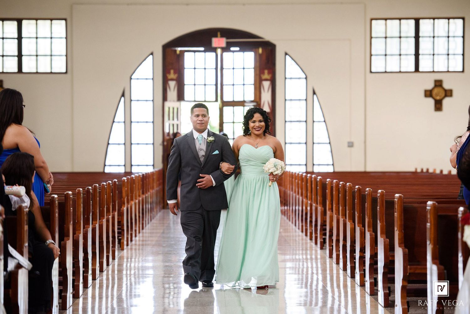 Bride in mint green gown walks down church aisle with escort during elegant wedding ceremony.