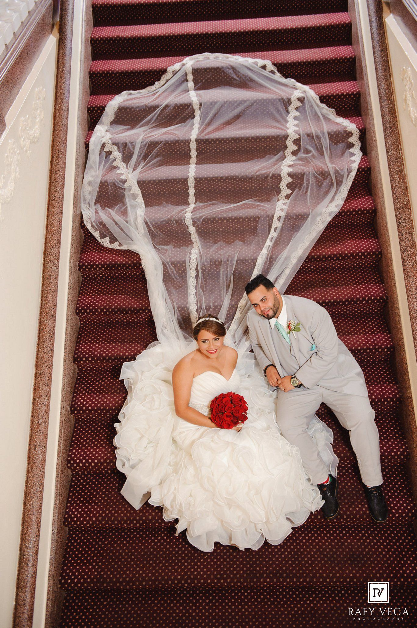 Aerial view of bride in white ruffled gown with flowing veil and groom in gray suit sitting on red carpeted stairs.