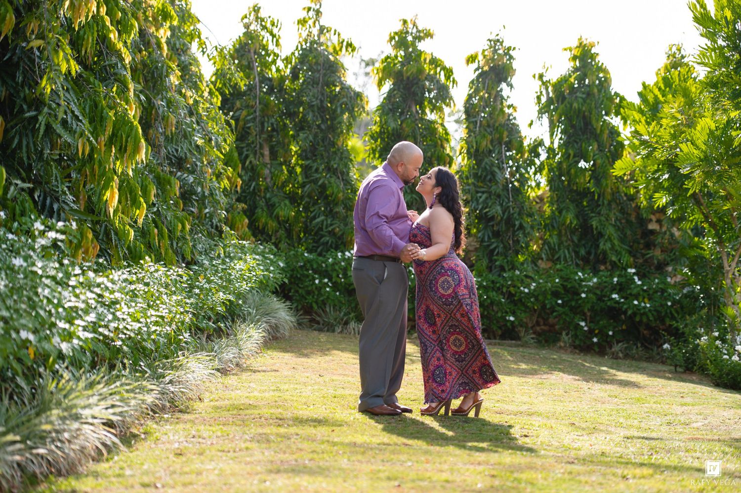 Couple sharing a romantic moment in a lush green garden surrounded by tall trees during golden hour.