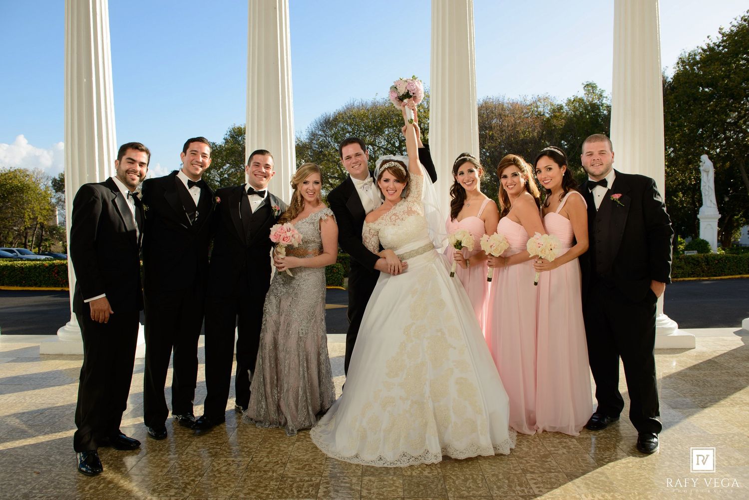 Wedding party poses with bride and groom between white columns, bridesmaids in pink gowns and groomsmen in black tuxedos.