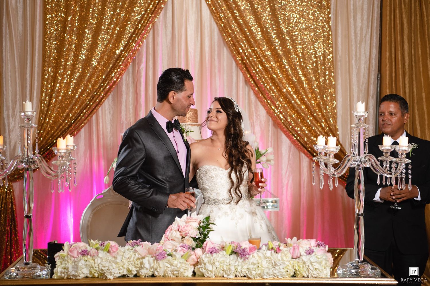 Bride and groom share a kiss at elegant wedding reception with gold sequin backdrop and floral arrangements.