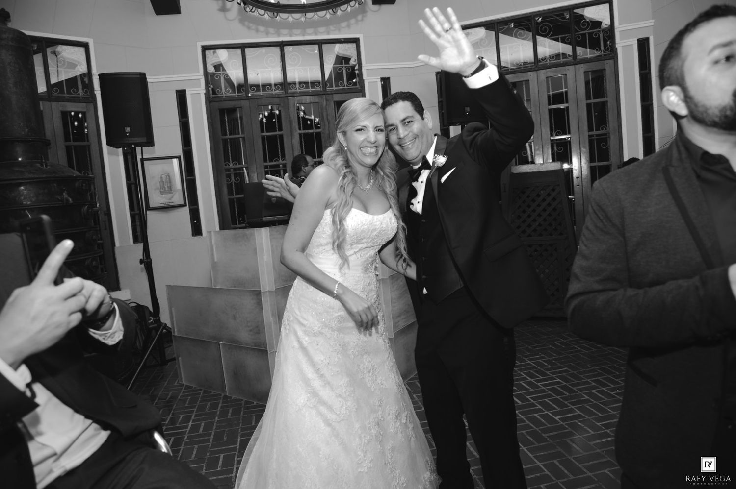 Black and white photo of smiling bride and groom waving at wedding reception celebration.