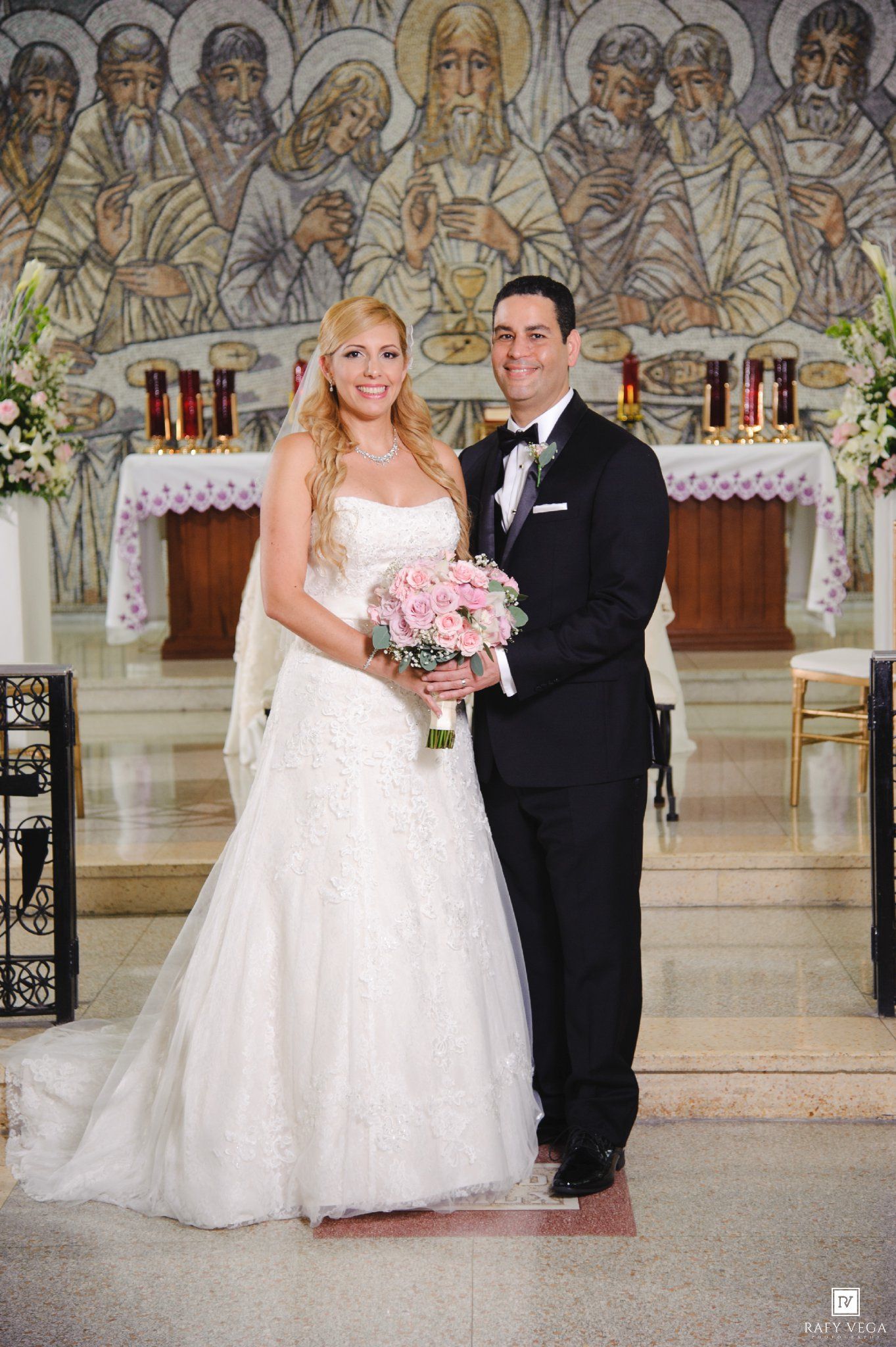 Bride and groom posing together at church altar, bride in white gown holding pink bouquet, groom in black tuxedo.